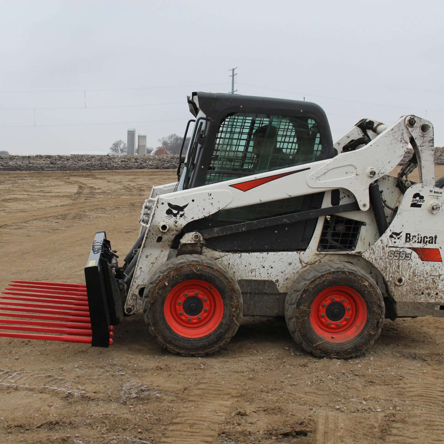 Side view of a skid steer loader equipped with Messer Attachments 84-inch heavy-duty manure fork on a dirt field.