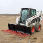 Angled front view of a skid steer loader fitted with Messer Attachments 84-inch manure fork featuring red spring steel tines