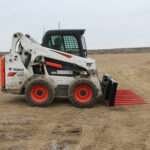 Side view of a skid steer loader equipped with Messer Attachments 84-inch heavy-duty manure fork on a dirt field