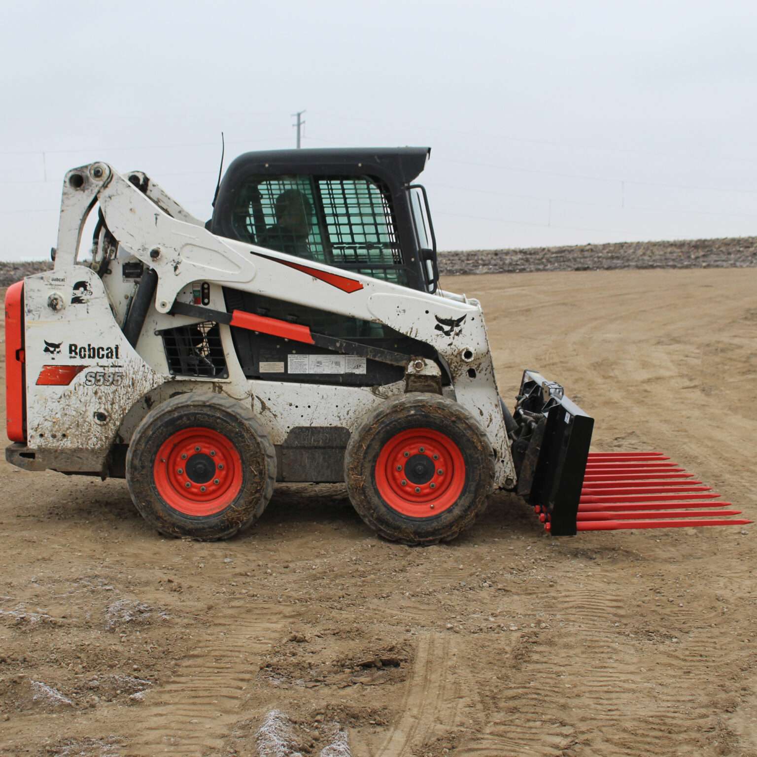 Side view of a skid steer loader equipped with Messer Attachments 84-inch heavy-duty manure fork on a dirt field