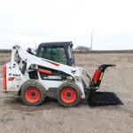 A Bobcat skid-steer loader with a Messer brush attachment and an open grapple is sitting on the ground. The Brush & Root Grapple has a maximum jaw opening of 42 inches. Messer Attachments